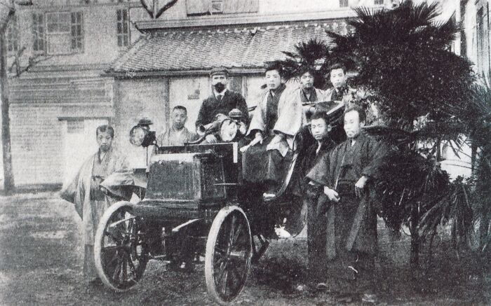 Early black and white photo showing a wild first car from 100 years ago with a group of people posing around it.