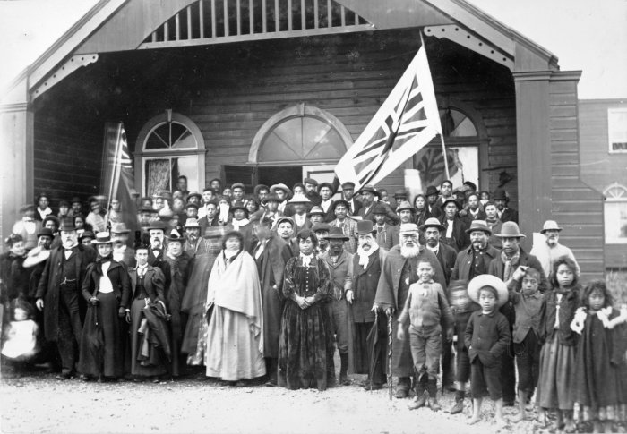 Large historical group portrait of Māori people, men, women, and children, gathered outside a wooden building with flags.