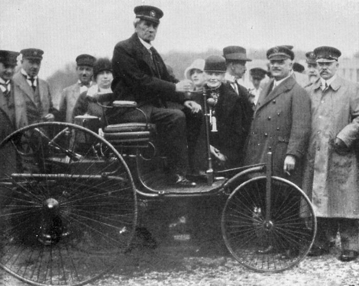 Early car with large spoked wheels and driver in uniform surrounded by a crowd, showing how wild the first cars really were.