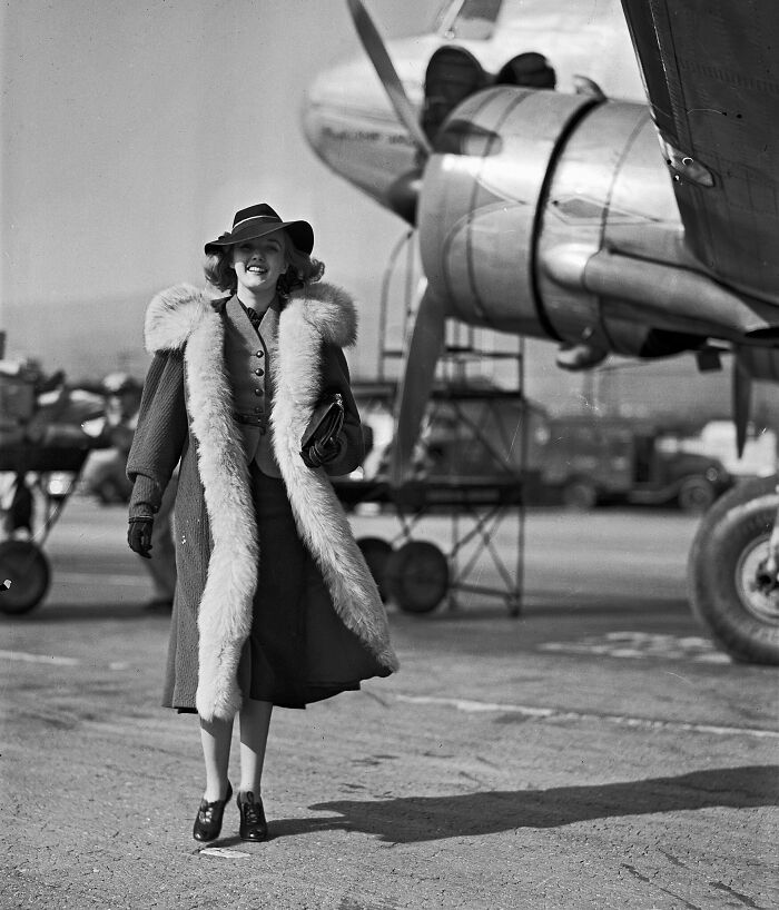 Woman in a fur-trimmed coat and hat standing by vintage airplane, reflecting flying glamour and style from a bygone era.