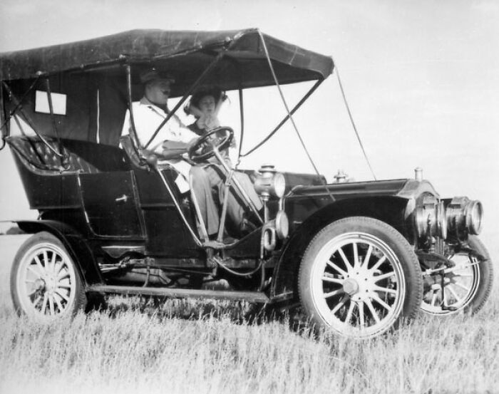 Vintage black and white photo showing a wild first car from 100 years ago with two people sitting inside.