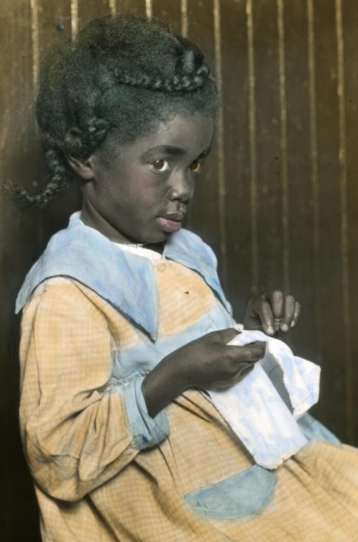 Colorized photo of an African American child in the 1920s, wearing a yellow dress and sewing fabric indoors.