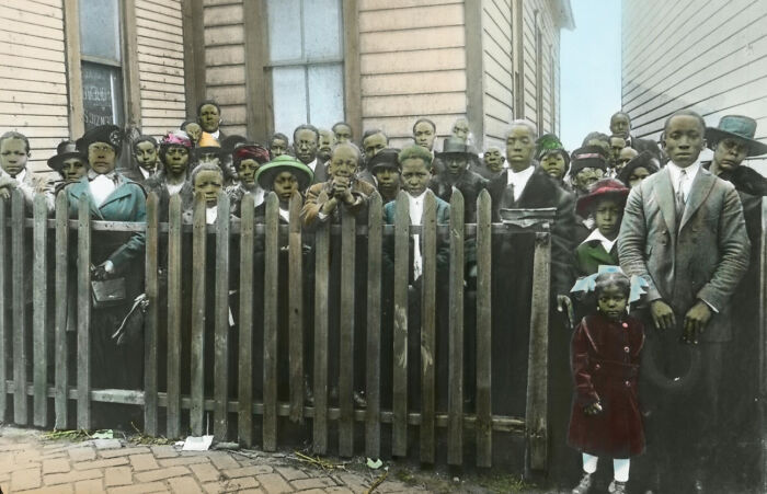 Group of African American men, women, and children from the 1920s standing behind a wooden fence in a colorized photo.