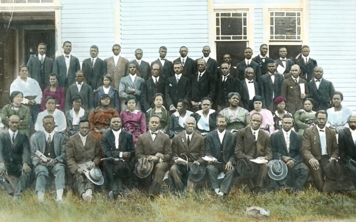Large group of African American men and women in formal 1920s attire posing outside a building, colorized photo showcasing history.