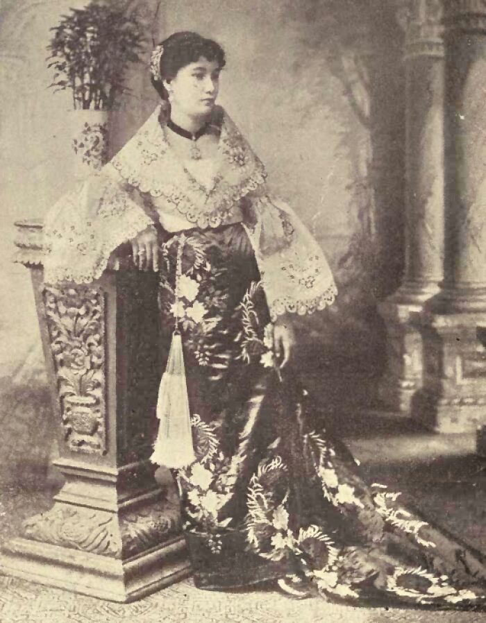 Filipina woman in traditional 1890s attire standing by an ornate pillar, showcasing fashion in the Philippines 1890s.