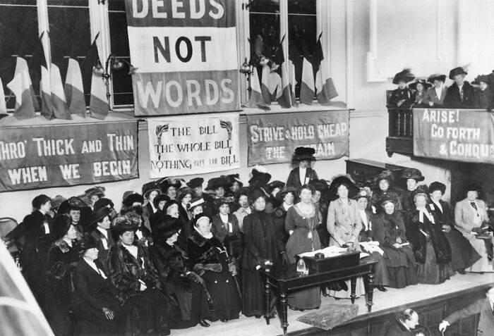 Black and white photo of women in the 20th century suffrage era gathered in a hall with banners advocating for voting rights.