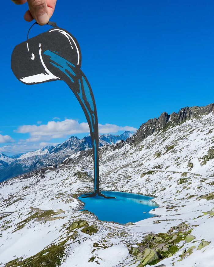 Hand holding paper cutout pouring painted water into lake, part of artist travels the world with paper cutouts project.