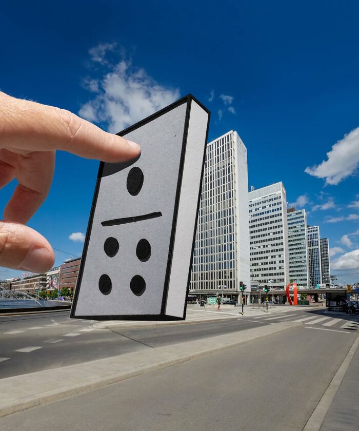 Hand holding a paper cutout domino over a city street, showcasing artist’s creative paper cutouts and surprising scenes.