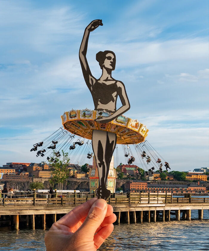 Hand holding a paper cutout of a ballerina positioned to blend with a swing carousel, showcasing artist travels the world.