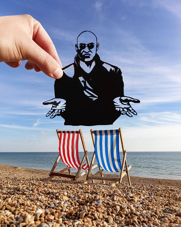 Hand holding a paper cutout of a seated figure above two striped beach chairs on a pebbled shore, art scene.