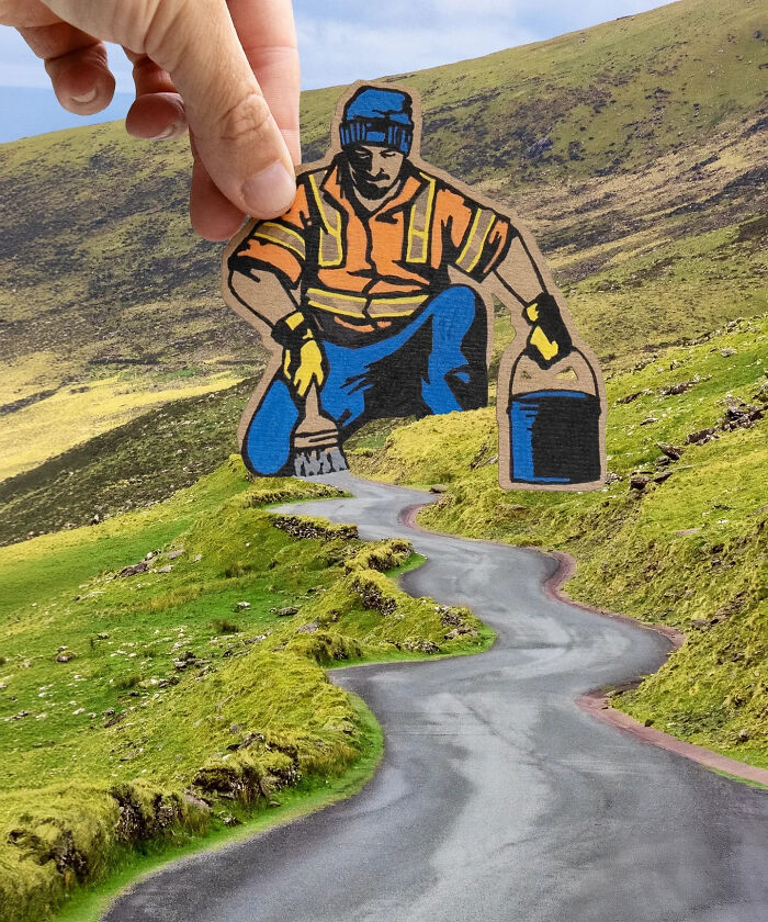 Hand holding a paper cutout of a painter working on a winding road in green hills, showcasing creative artist travels the world scenes.