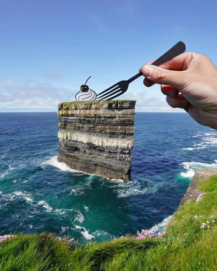 Hand holding a paper cutout of a fork with a dessert scene on a sea cliff, showcasing artist's creative travel art.