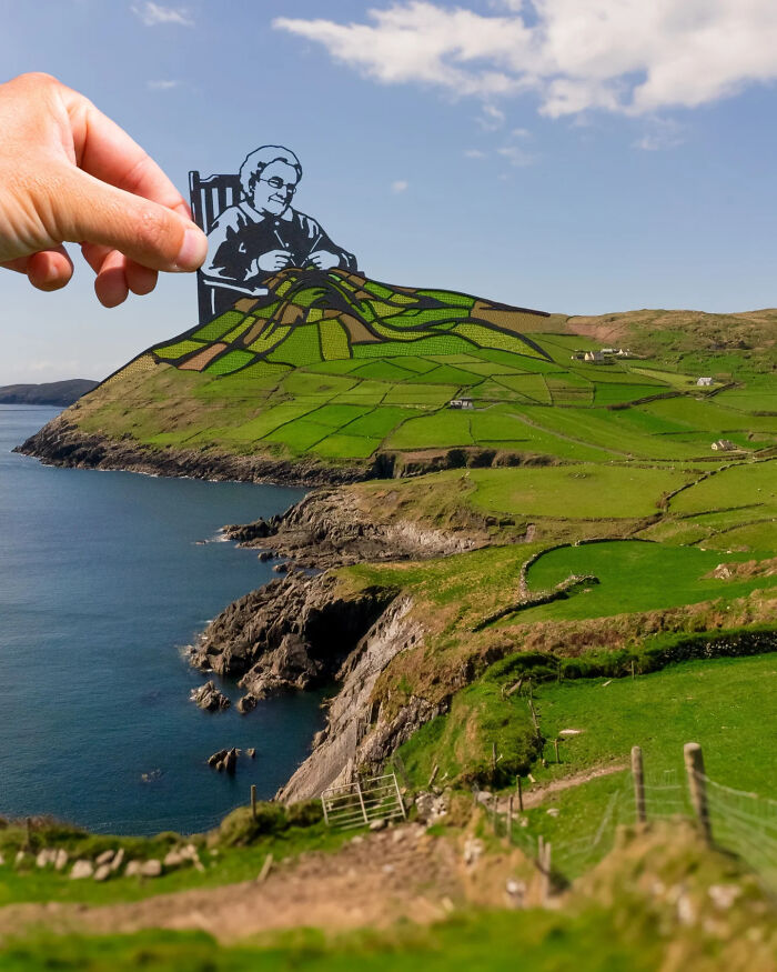 Hand holding paper cutout of an elderly woman knitting that blends into a green landscape scene by the coast.