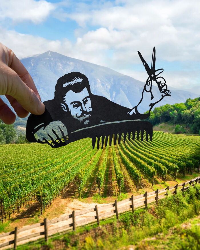 Hand holding a paper cutout of a man with scissors and a comb over a vineyard, artist travels the world with paper cutouts.