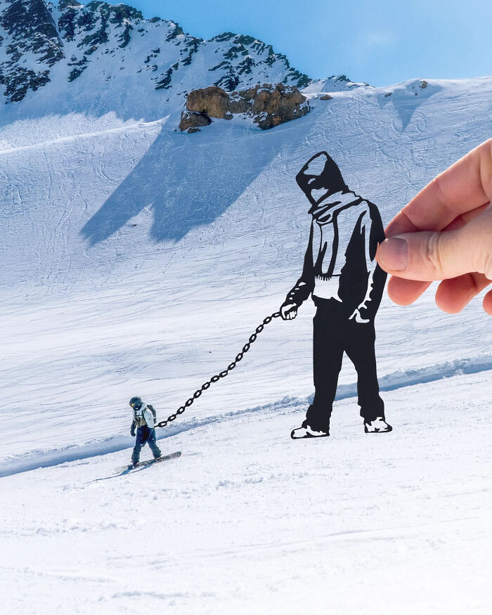 Hand holding paper cutout leash attached to skier on snow, artist travels the world creating surprising scenes.