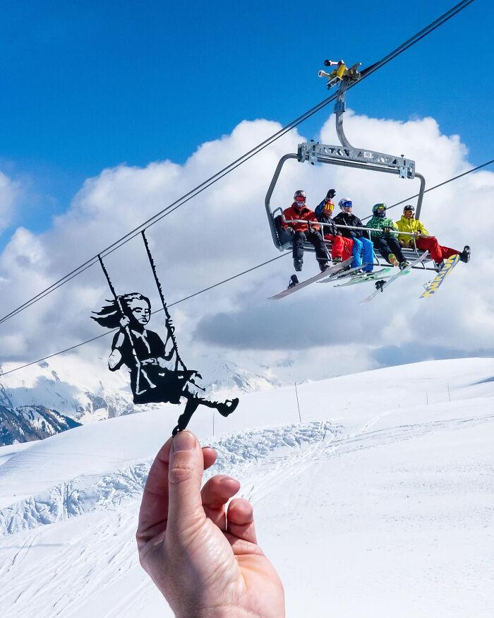 Hand holding paper cutout of a girl on a swing, aligned with skiers on a ski lift in snowy mountain scene.