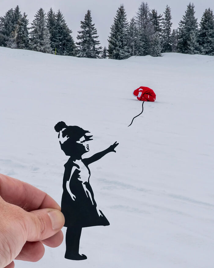 Hand holding a paper cutout of a girl reaching out to a red balloon on a snowy landscape, creative artist travels.