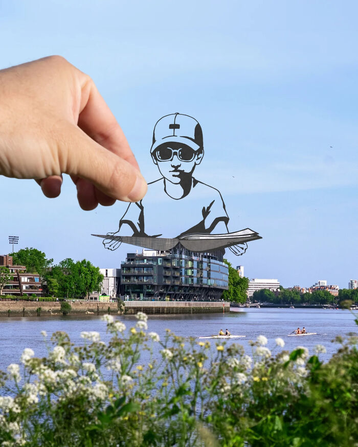 Hand holding paper cutout of a boy reading a book over a river scene, showcasing artist’s paper cutouts world travels.