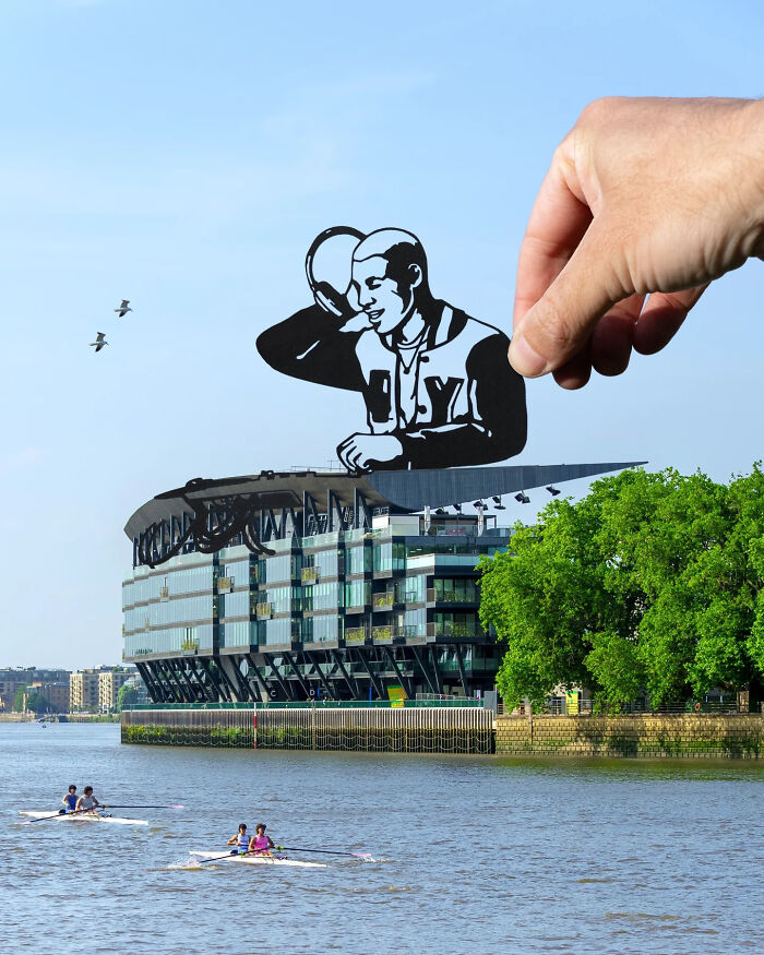 Hand holding paper cutout of two people whispering placed over river scene, artist travels the world with paper cutouts.
