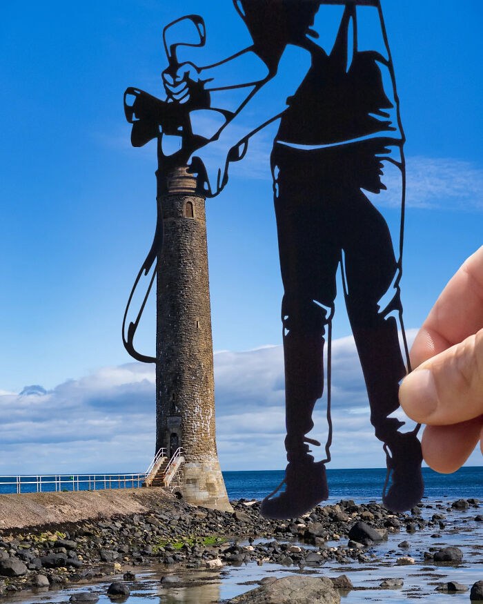 Hand holding paper cutout of a golfer aligned with a lighthouse, creating a surprising scene in an outdoor coastal setting.