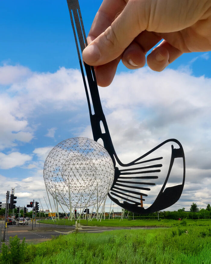 Hand holding paper cutout of golf club and ball aligned with large metal ball sculpture in outdoor scene, showcasing artist travels.