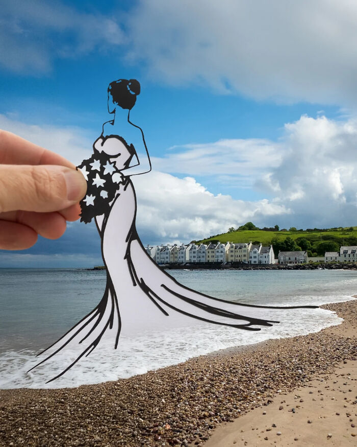Hand holding paper cutout of a woman in a dress blending with the beach and ocean waves, artist travels the world.