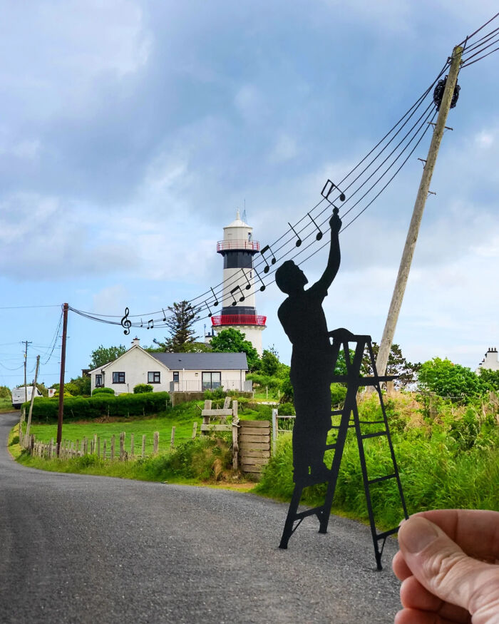 Paper cutout art shows a silhouette climbing a ladder to paint musical notes on wires in a countryside scene.
