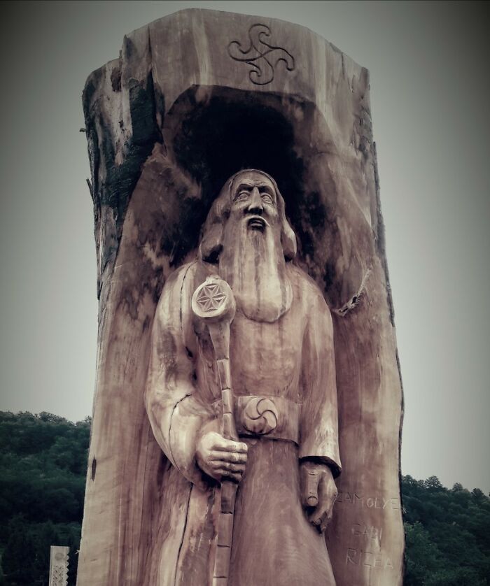 Chainsaw artist carved a detailed wooden statue of a bearded figure holding a staff from a large forgotten tree trunk outdoors.