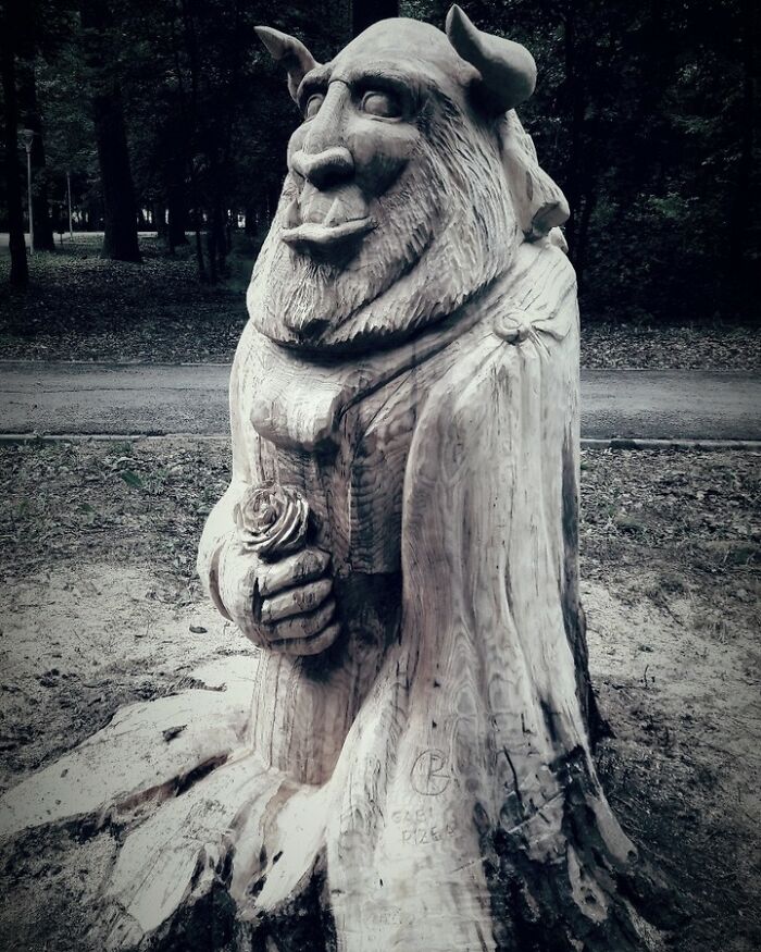 Chainsaw artist transforms a tree stump into detailed art sculpture of a mythical creature holding a rose in a forest setting.