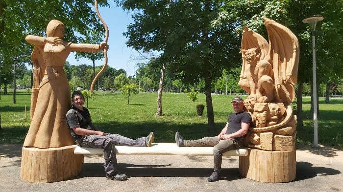 Two men sitting on a bench carved from a tree, featuring chainsaw art sculptures of an archer and a dragon.