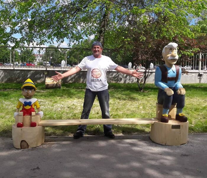 Chainsaw artist standing between colorful wooden sculptures of Pinocchio and an old man bench outdoors under a tree.