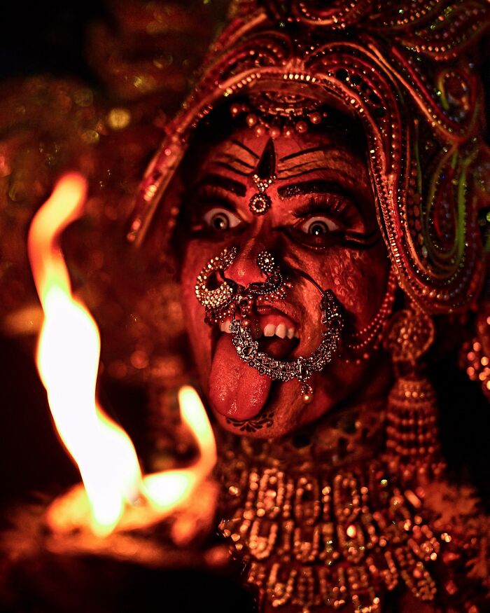 Close-up of a vibrant performer in traditional Indian attire with elaborate jewelry and face paint near a bright flame.