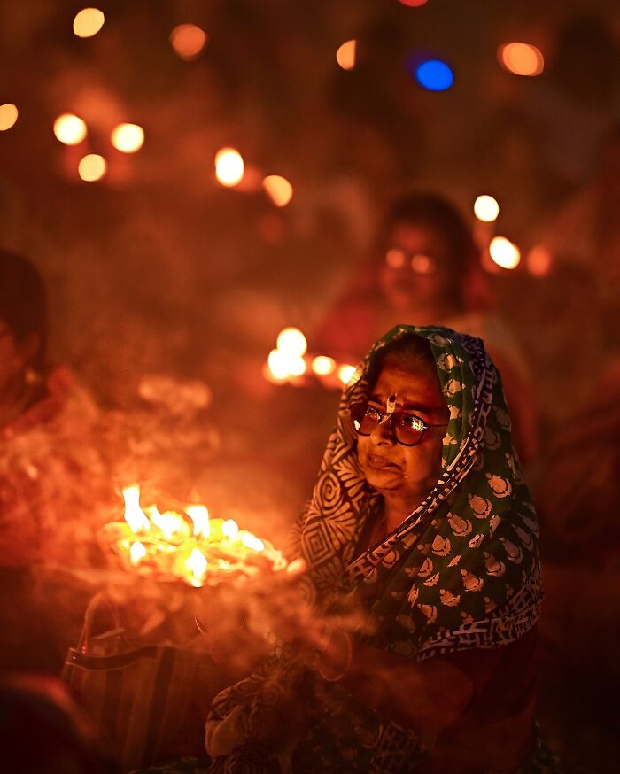Elderly woman wearing a patterned saree holding lit candles during a colorful Indian festival at night bursting with vibrant life.