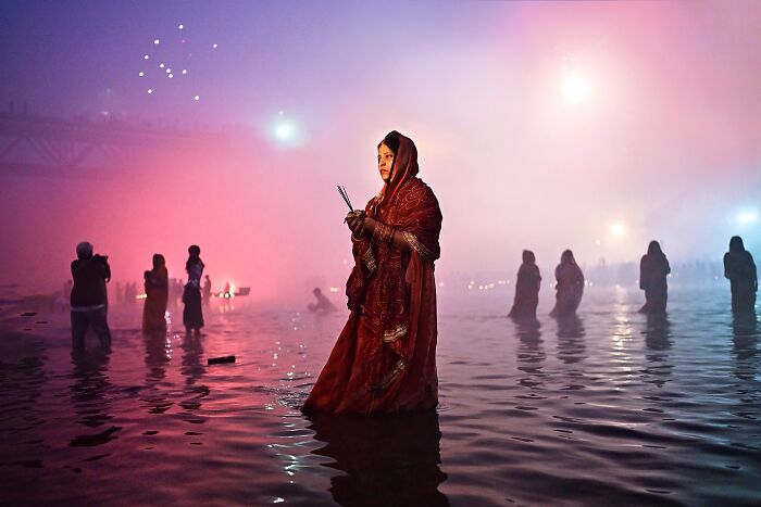 Woman in a vibrant red sari standing in water during a colorful Indian festival filled with lights and mist.
