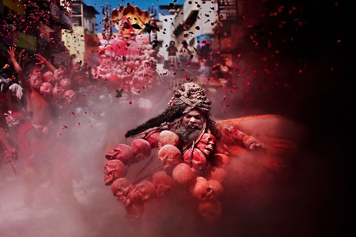 A vibrant scene in India showing a man adorned with skull necklaces amidst colorful powder and festive crowd.