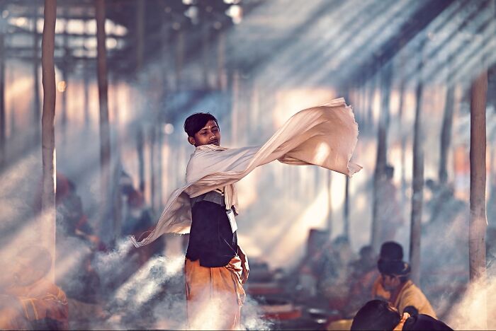 Young Indian man swirling fabric in a sunlit, smoky environment, showcasing vivid colors and life in India photography.
