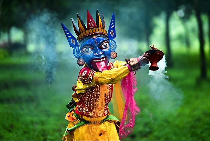 Colorful traditional Indian dancer in vibrant costume and mask performing ritual with incense in lush green setting