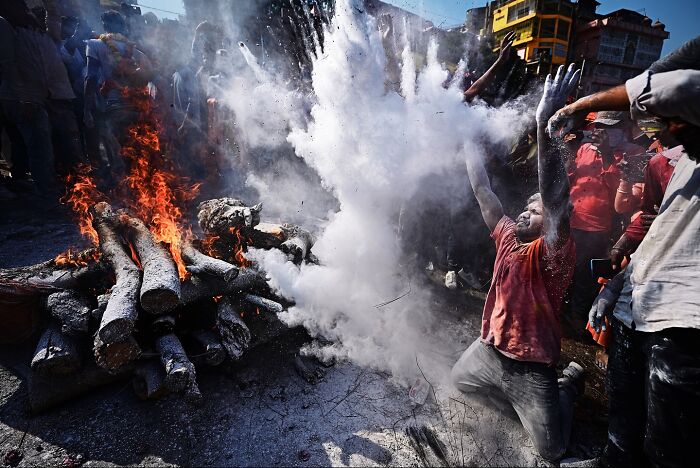 Man covered in white powder raising arms near burning logs during vibrant festival in India bursting with color and life