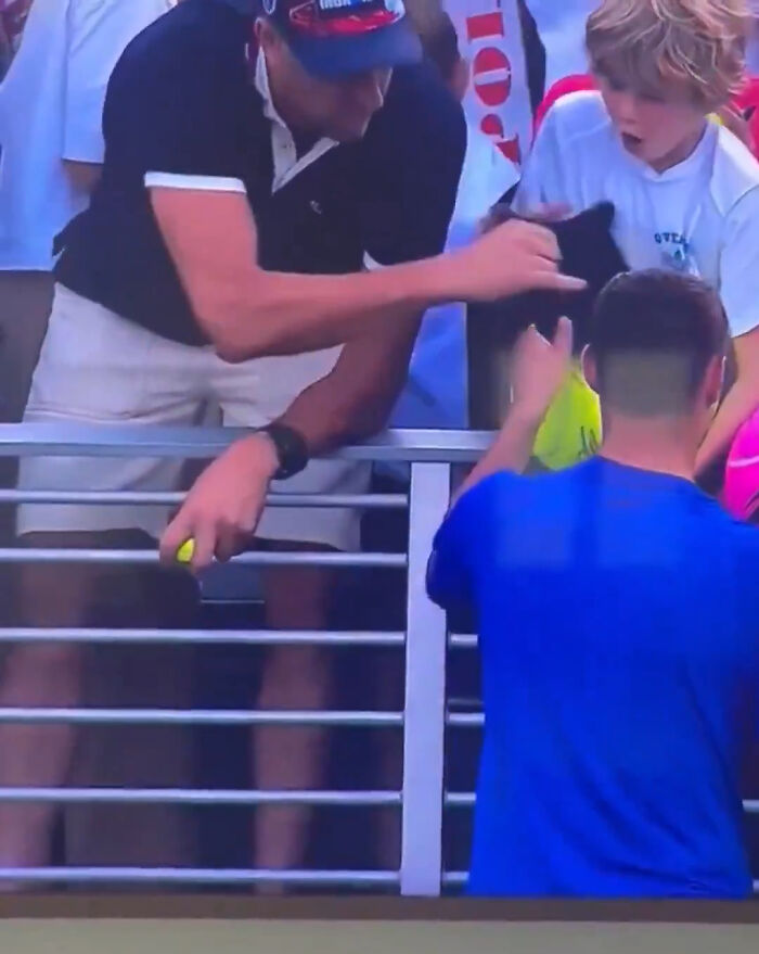 Polish CEO reaching over railing to grab a hat during US Open, with surprised child and other spectators nearby. Polish CEO reaching over railing to grab a hat during US Open, with surprised child and other spectators nearby.