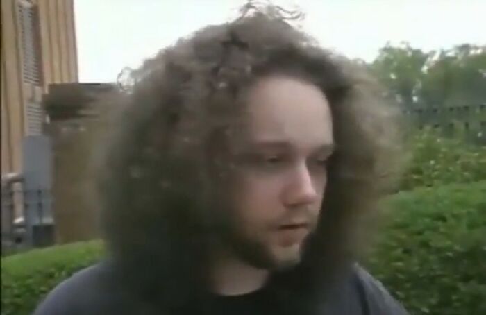 Young man with curly hair standing outdoors, looking down thoughtfully in a video with hidden context.