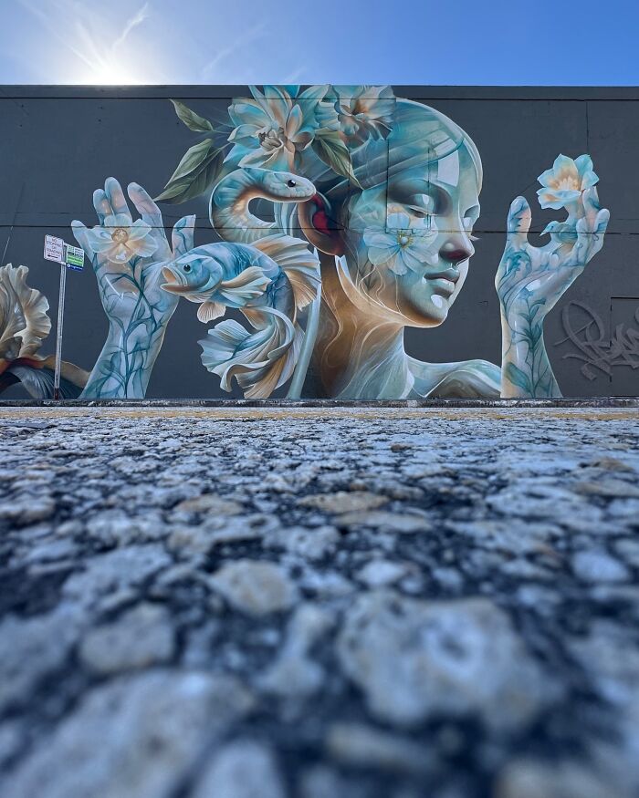 Large mural blending graffiti and realism depicting a serene woman with flowers, fish, and a snake on a building wall.
