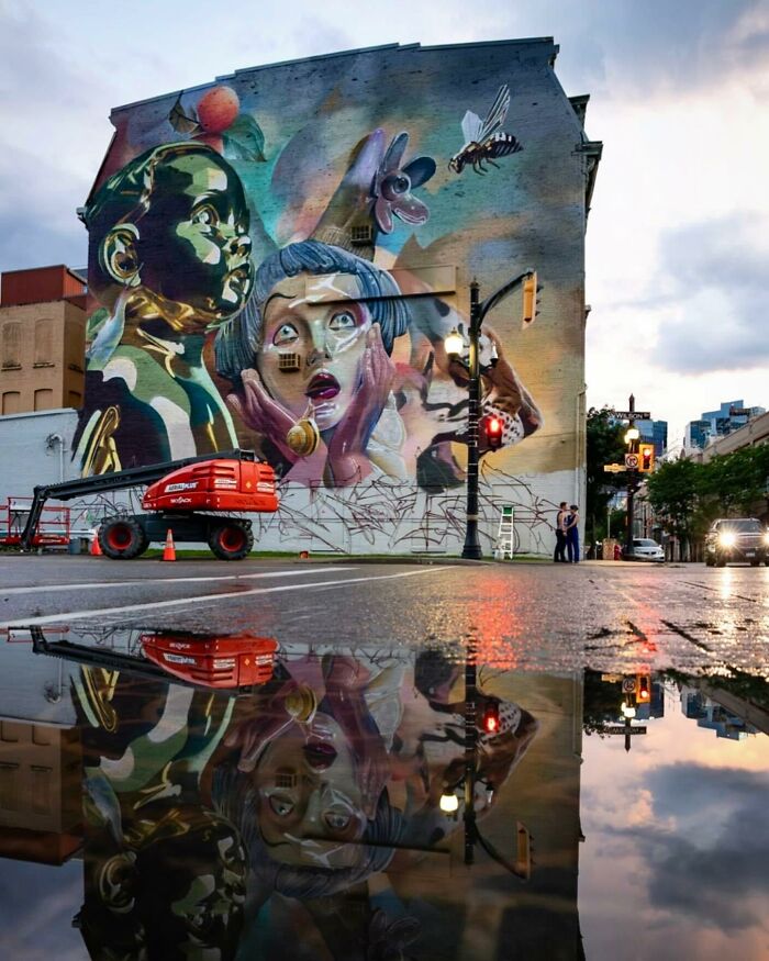 Large mural blending graffiti, realism, and abstraction on a building wall, reflected in a wet street puddle.