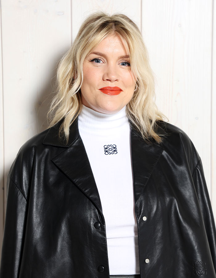 Blonde woman wearing black leather jacket and white turtleneck, posing against light wooden wall for Wuthering Heights adaptation. Blonde woman wearing black leather jacket and white turtleneck, posing against light wooden wall for Wuthering Heights adaptation.