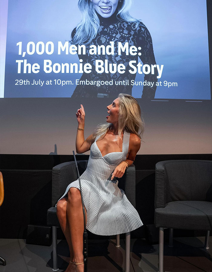 Woman in a white dress seated on stage, pointing up at a screen about Bonnie Blue’s career in a new documentary. Woman in a white dress seated on stage, pointing up at a screen about Bonnie Blue’s career in a new documentary.