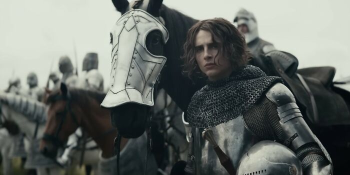 Young knight in medieval armor standing next to a horse with a decorative metal faceplate in a Netflix movies scene