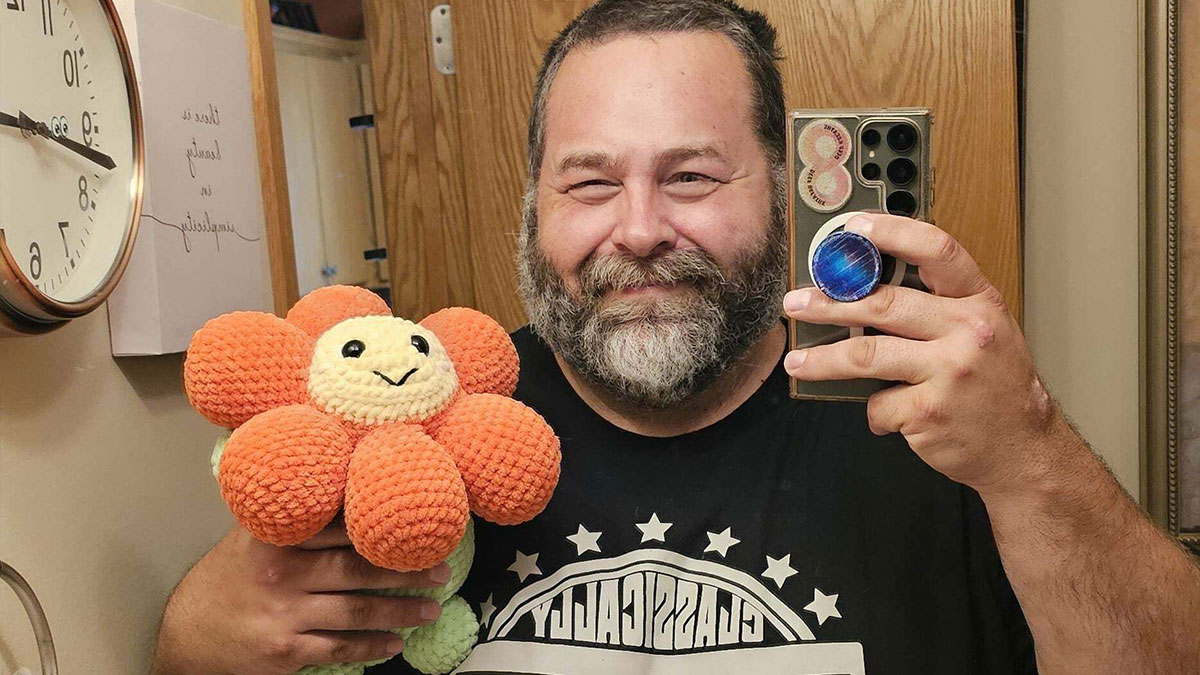 Man smiling and holding a crocheted plush flower, taking a mirror selfie showcasing crochet craftsmanship.