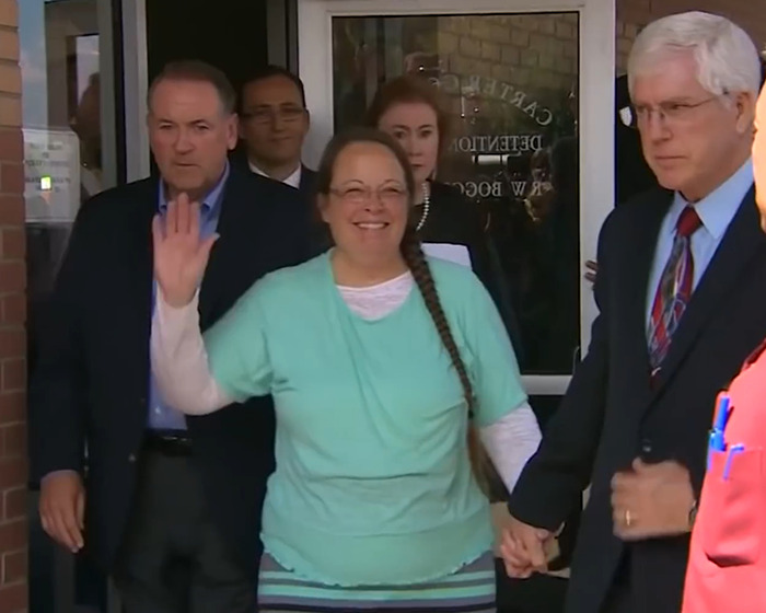 Kim Davis smiling and waving while leaving a courthouse, surrounded by several people including a man in a suit holding her hand. Kim Davis smiling and waving while leaving a courthouse, surrounded by several people including a man in a suit holding her hand.