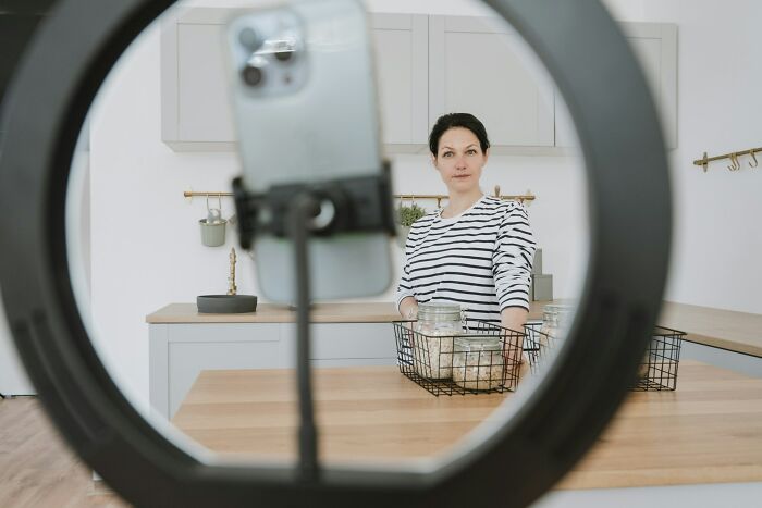 Woman recording a video in a kitchen using a smartphone and ring light, demonstrating content for viewers with context.