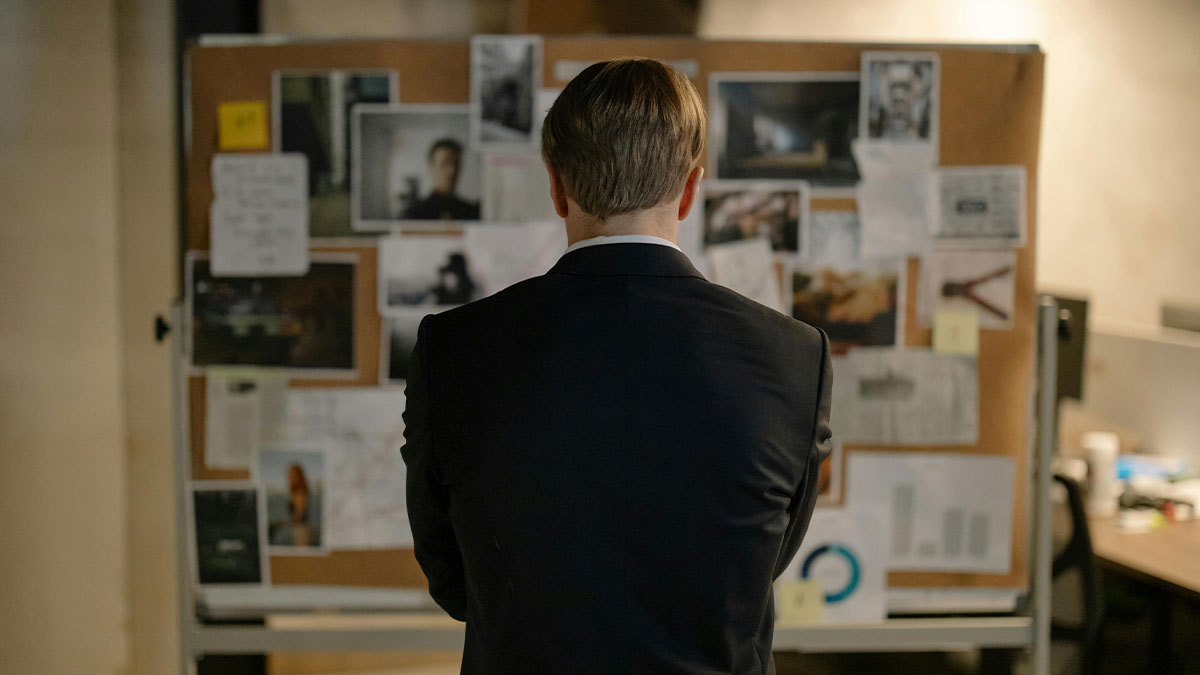 Retired major crimes detective in suit analyzing case details on a cluttered evidence board in an office setting.