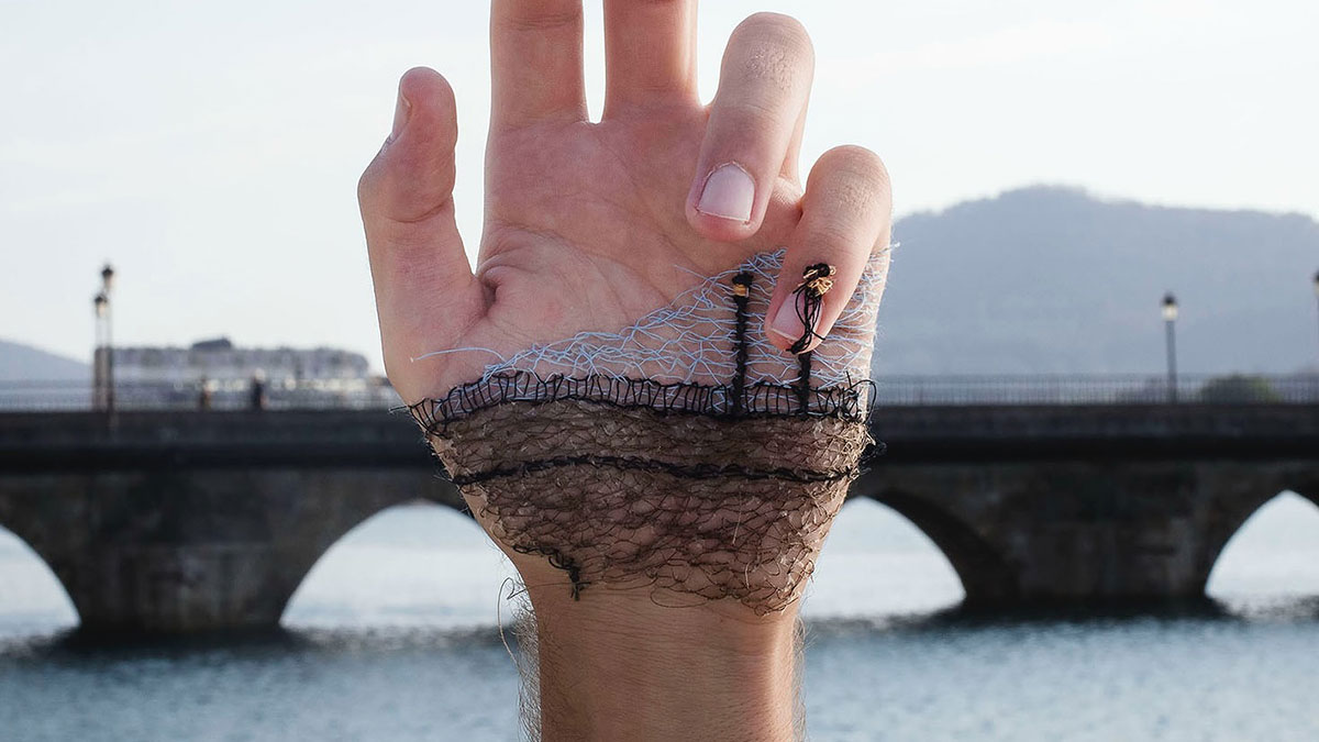 Close-up of a hand with stitched landscape art, blending threads to create a bridge and water scene on the skin.